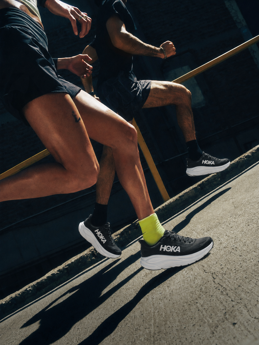 Runners wearing black HOKA sneakers with white cushioned soles while running outdoors in athletic clothing.
