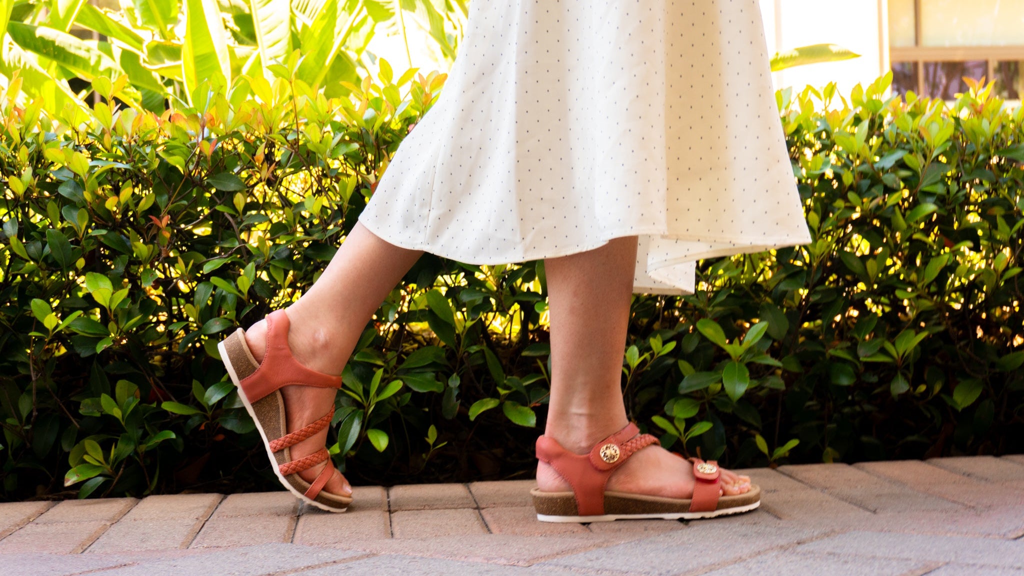 A person wearing terracotta-colored strappy sandals with a white skirt, walking outdoors on a paved path with green hedges in the background.