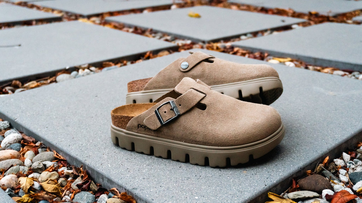 A pair of taupe-brown clog-style shoes with a single strap and silver buckle, resting on gray square patio stones outdoors with pebbles and fallen leaves around them.