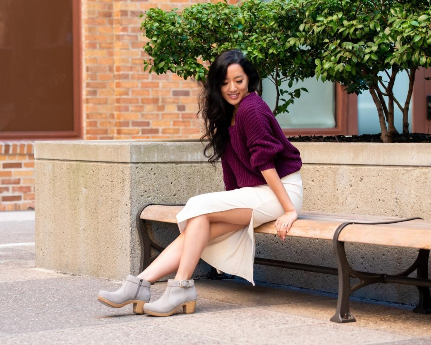 A woman sits on a concrete bench outdoors, wearing a dark purple knit cardigan, a white midi skirt, and light gray heeled ankle boots. The background features a brick building, a concrete planter with green shrubs, and a paved walkway.